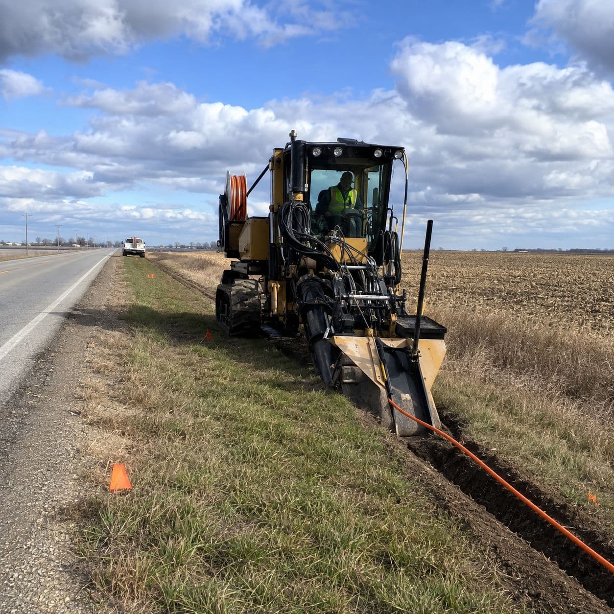 A tracked cable plow pulling orange conduit into the ground along a rural highway right-of-way, the operator in a hi-vis vest at the controls, a conduit reel mounted behind the cab, and a narrow spoil line and locate flags trailing back down the shoulder