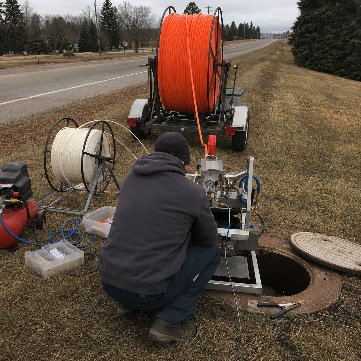 A technician kneeling at a fiber jetting machine set over an open roadside vault, feeding cable into the duct below, with a trailer-mounted reel of orange conduit, a white fiber reel, an air compressor, and hand tools laid out on the shoulder beside a rural highway